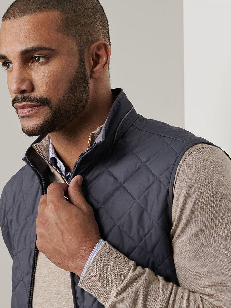 A man wearing the Peter Millar Essex Vest in Iron, layered over a beige sweater, looks to the side while holding the vest's zipper.