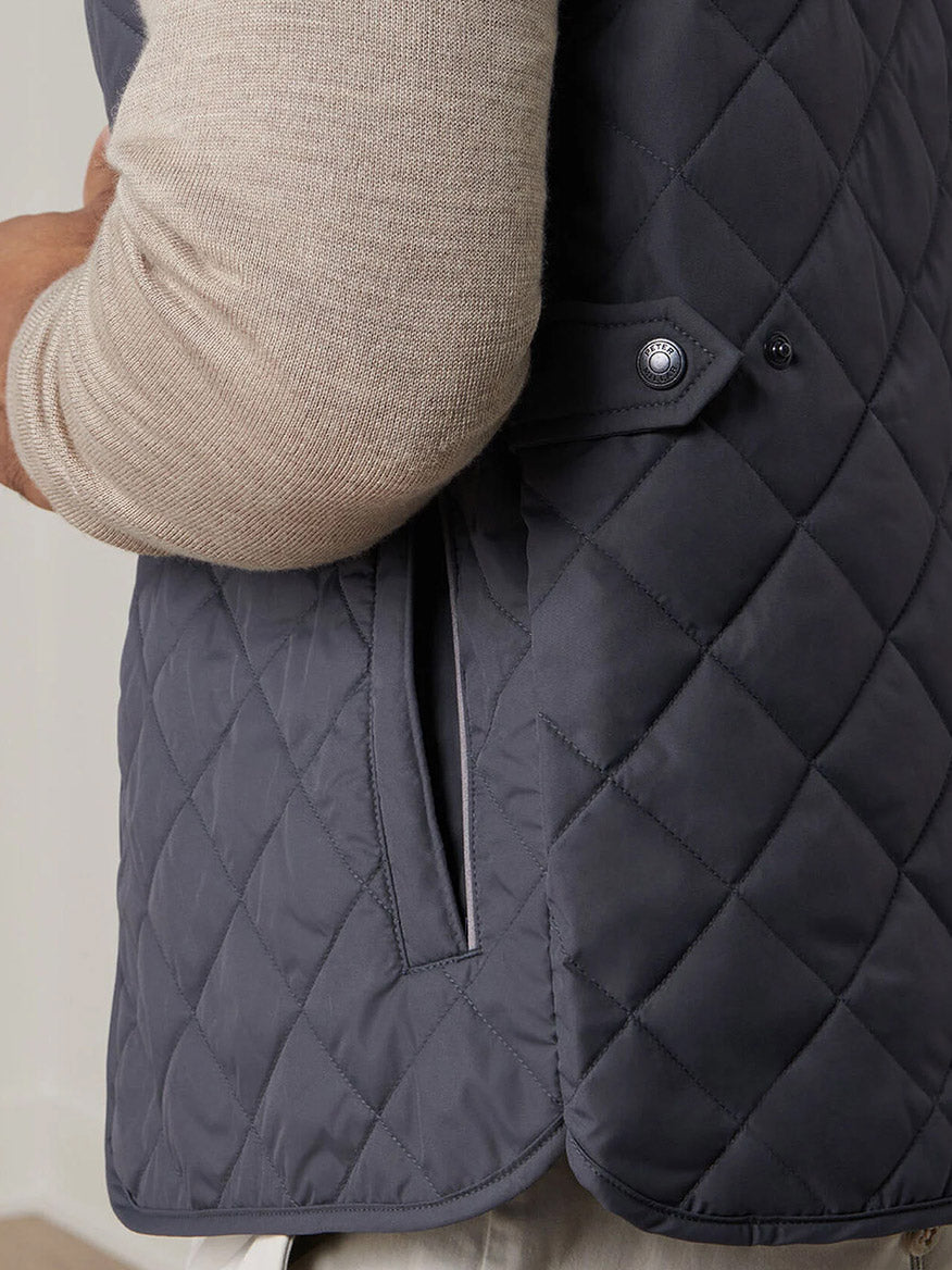 A person wearing a beige long-sleeve shirt and the Peter Millar Essex Vest in Iron—a quilted, dark gray windproof vest featuring a side pocket with buttoned flap detail.
