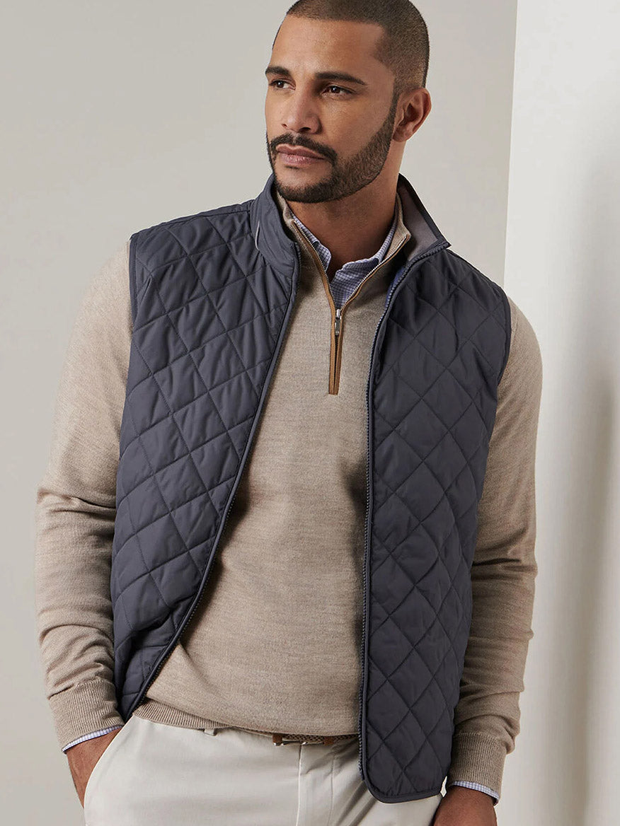 A man with short hair and a beard wears a beige sweater, light pants, and the Peter Millar Essex Vest in Iron, standing against a plain light background.