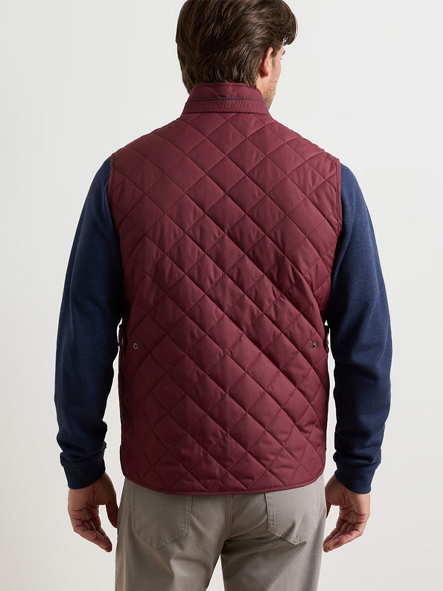 A man is shown from behind wearing the Peter Millar Essex Vest in Bordeaux over a navy long-sleeve shirt and khaki pants, standing against a plain light background.