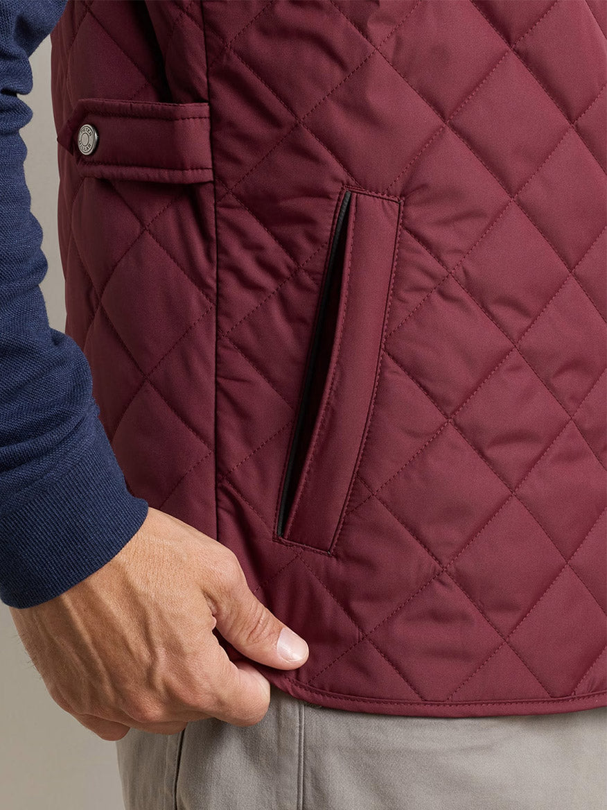 A person wearing the Peter Millar Essex Vest in Bordeaux over a navy long-sleeve shirt and beige pants, with their hand resting near the vest pocket.