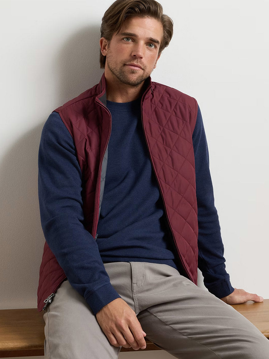 A man with light brown hair wears the Peter Millar Essex Vest in Bordeaux, a water-resistant quilted vest, over a navy sweater and light gray pants as he sits on a wooden surface against a plain background.