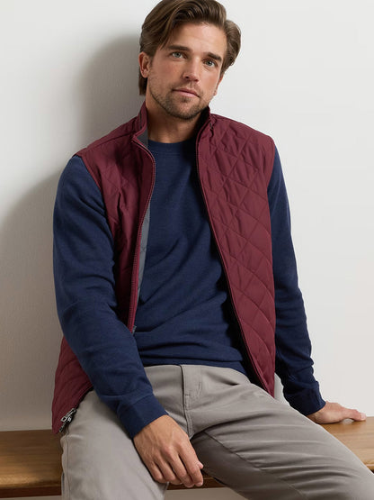 A man with light brown hair wears the Peter Millar Essex Vest in Bordeaux, a water-resistant quilted vest, over a navy sweater and light gray pants as he sits on a wooden surface against a plain background.