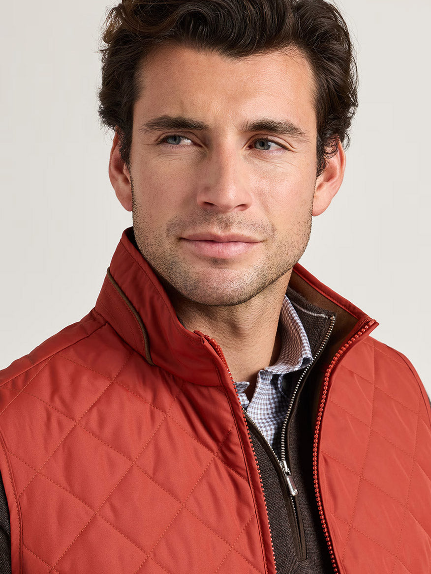 A man with short brown hair wears the Peter Millar Essex Vest in Burnt Orange—a windproof and water-resistant quilted layer—over a brown zip-up and checkered shirt, gazing slightly to the side against a plain light background.