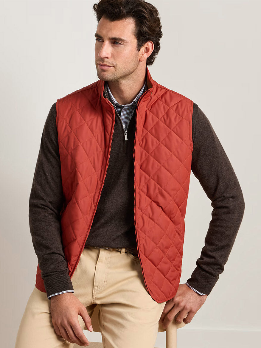 A man wearing a Peter Millar Essex Vest in Burnt Orange over a brown sweater and beige pants sits on a stool against a plain light background.