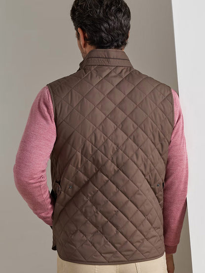 A man is seen from behind, standing near a white wall, wearing the Peter Millar Essex Vest in French Press—a water-resistant, quilted brown vest—over a pink long-sleeve shirt.