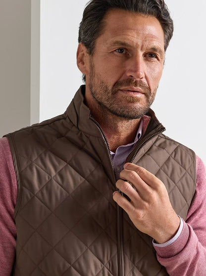 A man with short dark hair stands indoors, wearing the Peter Millar Essex Vest in French Press—a water-resistant quilted brown vest—over a pink shirt, holding the vest’s zipper.