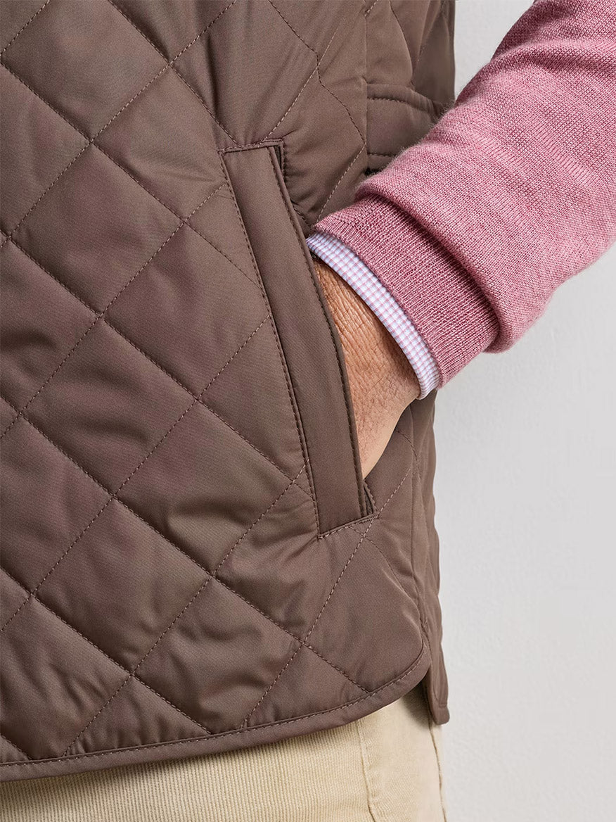 A person wearing the Peter Millar Essex Vest in French Press, layered over a pink sweater and paired with beige pants, stands with their hand in the vest pocket against a plain background.