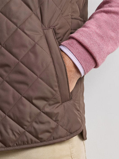 A person wearing the Peter Millar Essex Vest in French Press, layered over a pink sweater and paired with beige pants, stands with their hand in the vest pocket against a plain background.