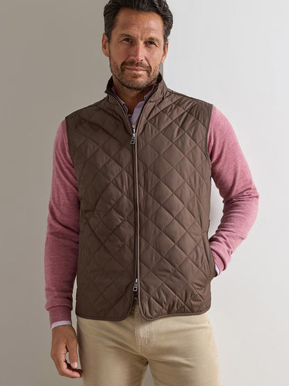 Man wearing the Peter Millar Essex Vest in French Press over a pink long-sleeve shirt and beige pants, standing against a plain light background.