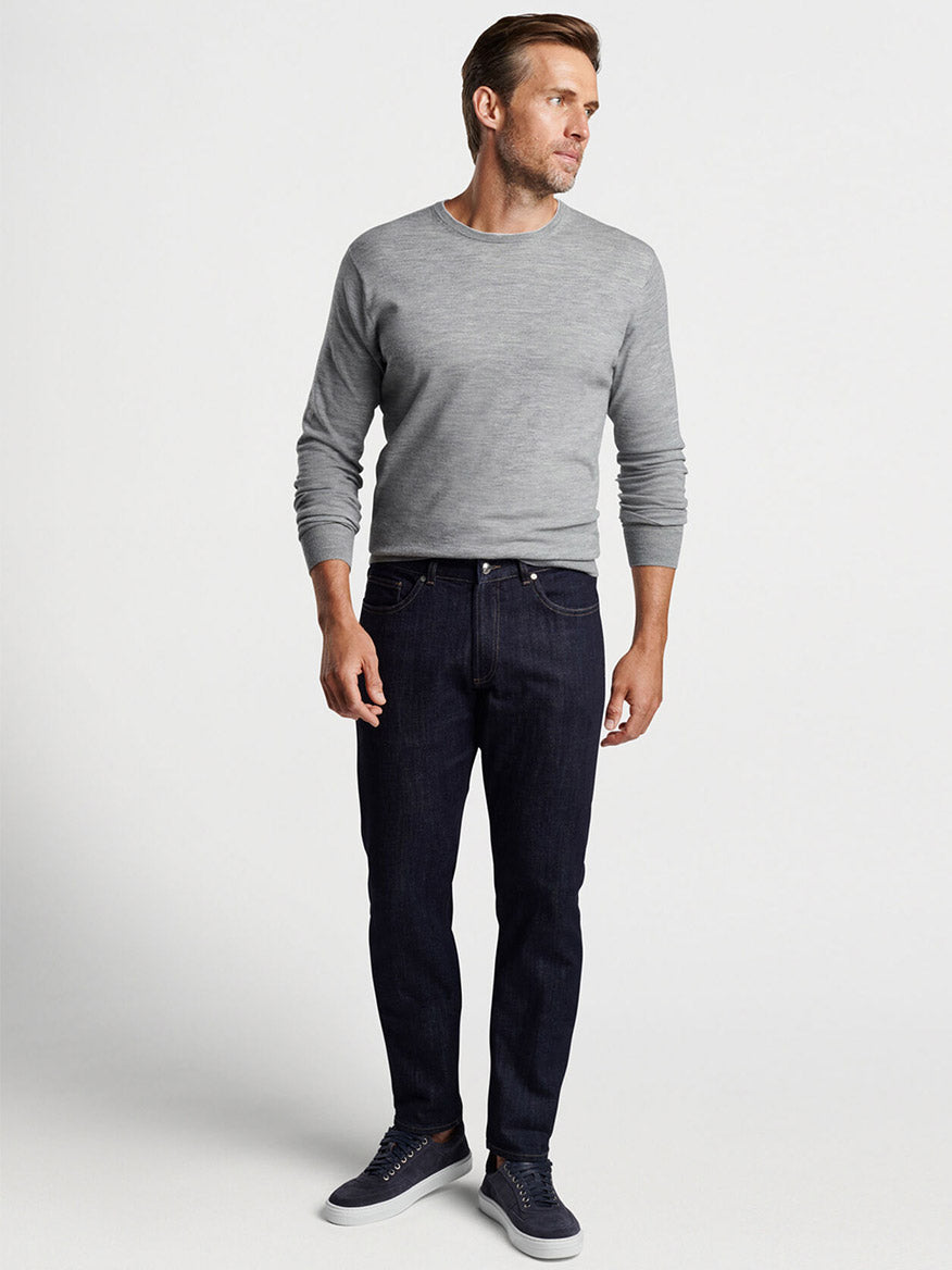 Man wearing a Peter Millar Excursionist Flex Crew in Gale Grey long-sleeve sweater, dark jeans, and blue sneakers, posing with hands in pockets.