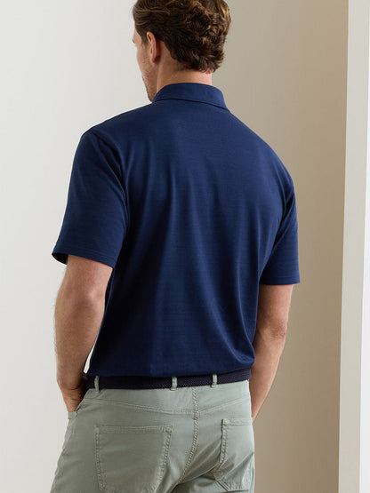 A man stands with his back to the camera, wearing a Peter Millar Excursionist Flex Herringbone Knit Polo in navy and light grey-green pants.