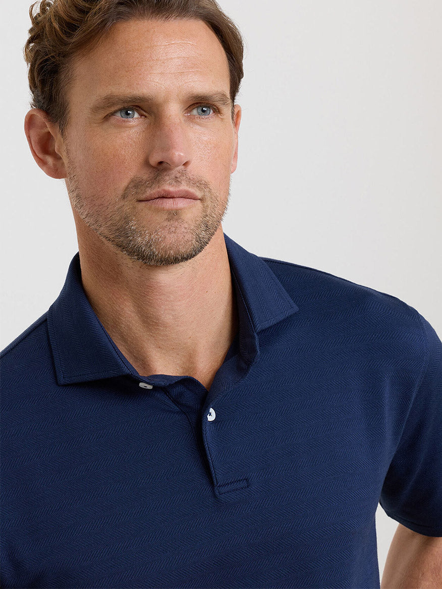 A man with short brown hair and a trimmed beard wears a navy Peter Millar Excursionist Flex Herringbone Knit Polo, looking slightly to the left against a plain light background.