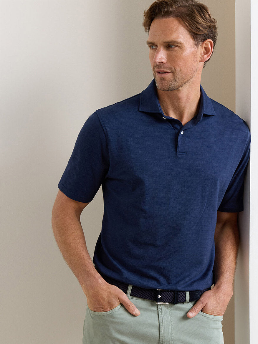 A man wearing a Peter Millar Excursionist Flex Herringbone Knit Polo in Navy and light khaki pants stands indoors, leaning against a wall with one hand in his pocket.