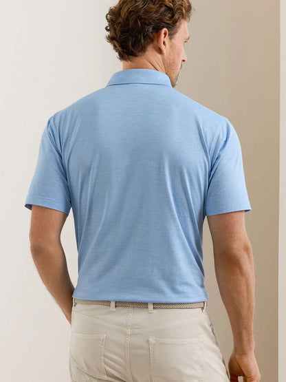 A man faces away in a tailored fit Peter Millar Excursionist Flex Polo in Blue Topaz and beige pants, standing before a neutral indoor background.