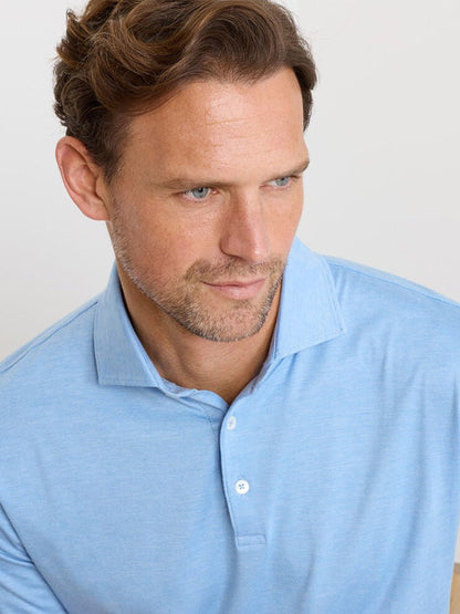Man with short brown hair and light stubble wears a Peter Millar Excursionist Flex Polo in Blue Topaz, made from cotton jersey knit, looking slightly down and to the side against a plain background.