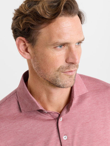 A man with light brown hair and stubble wears a Peter Millar Excursionist Flex Polo in Faded Scarlet, tailored fit, looking to the side against a plain background.