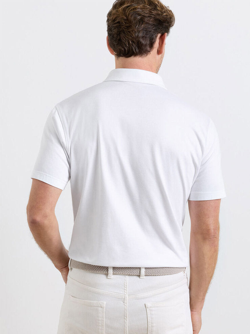 A man is shown from the back wearing the Peter Millar Excursionist Flex Polo in White, tailored fit, crafted from cotton-based jersey knit, tucked into cream-colored pants with a belt.