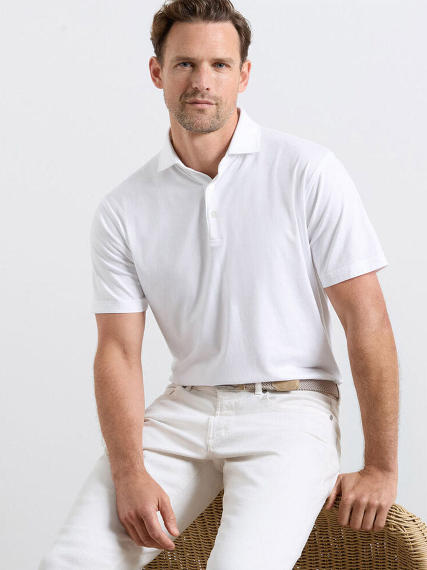 A man in a Peter Millar Excursionist Flex Polo in White and white pants sits on a woven chair against a plain light background.