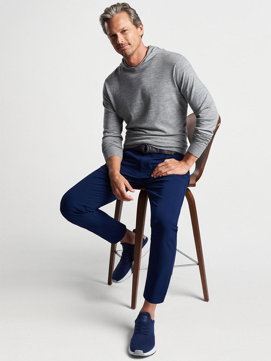 Man in a luxury Peter Millar Excursionist Flex Popover Hoodie in Gale Grey and blue trousers posing with a chair.