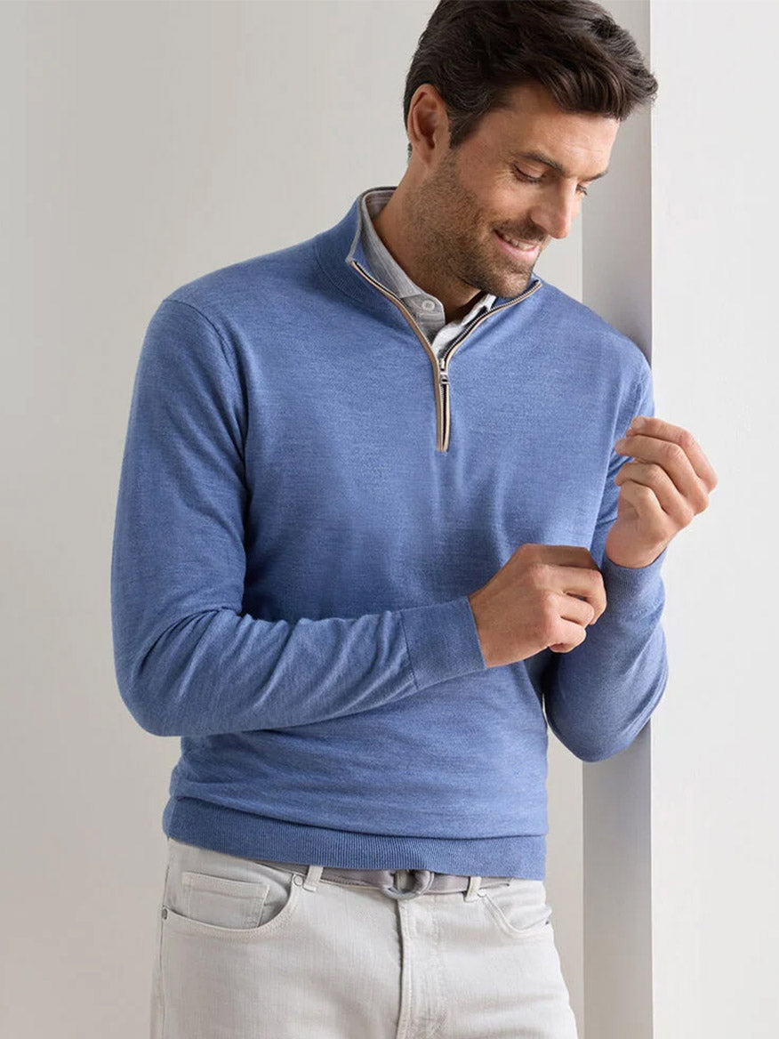 A man in a navy Peter Millar Excursionist Flex Quarter-Zip Sweater over a collared shirt stands against a white wall, looking down as he adjusts his sleeve.