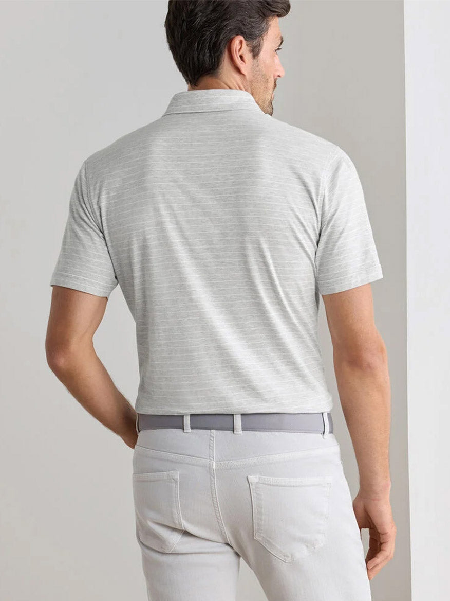 A man faces away, wearing a Peter Millar Excursionist Flex Striped Polo in British Grey, paired with matching gray pants and a gray belt, in a neutral indoor setting.