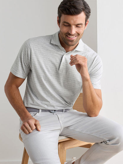 A man wearing a Peter Millar Excursionist Flex Striped Polo in British Grey and light gray pants sits on a wooden chair, smiling and looking down.