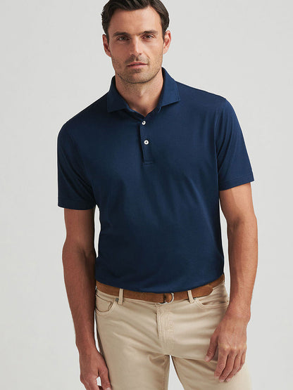 A man, dressed in a Peter Millar Excursionist Flex Polo in Atlantic Blue and beige pants, stands confidently against a plain background.