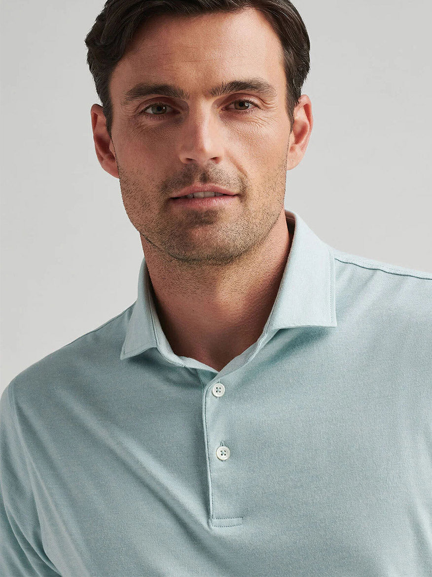 A man wearing a Peter Millar Excursionist Flex Polo in Blue Spruce jersey knit gazes at the camera with a neutral expression, highlighting the shirt's four-way stretch.