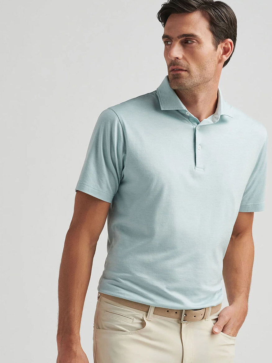 A man in a light blue Peter Millar Excursionist Flex Polo and beige pants stands against a plain background, looking to the side. The jersey knit is cotton-based with four-way stretch for optimal comfort and ease of movement.