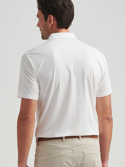 A person is wearing a Peter Millar Excursionist Flex Polo in White with beige pants, facing away from the camera against a plain background, highlighting the polo's tailored fit, sleek design, and four-way stretch comfort.