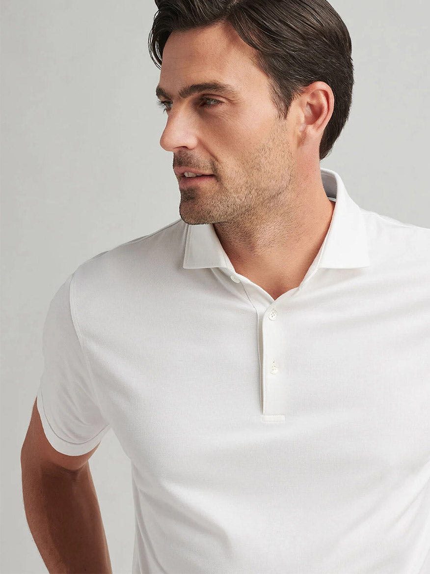 A man in a Peter Millar Excursionist Flex Polo in White, featuring a tailored fit and four-way stretch, gazes thoughtfully to the side against a plain background.