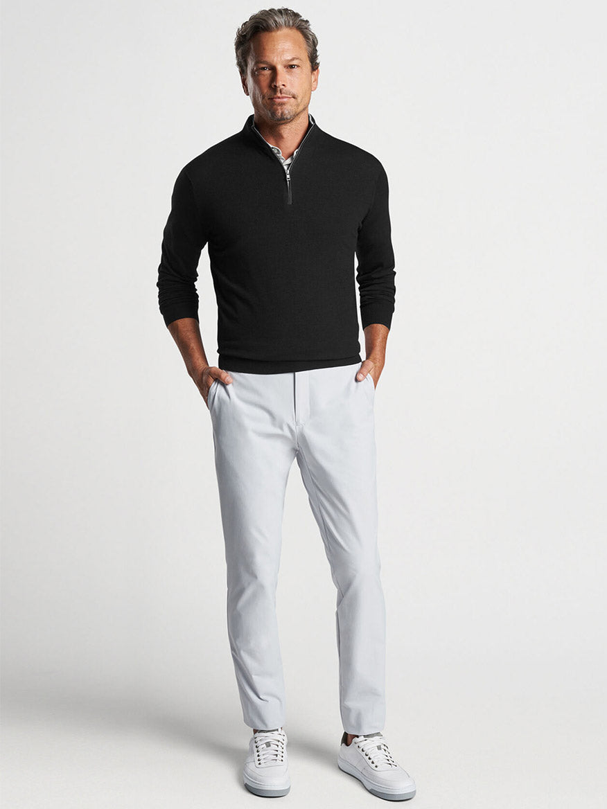 Man in Peter Millar Excursionist Flex Quarter-Zip in Black made of performance yarn and white trousers standing against a neutral background.