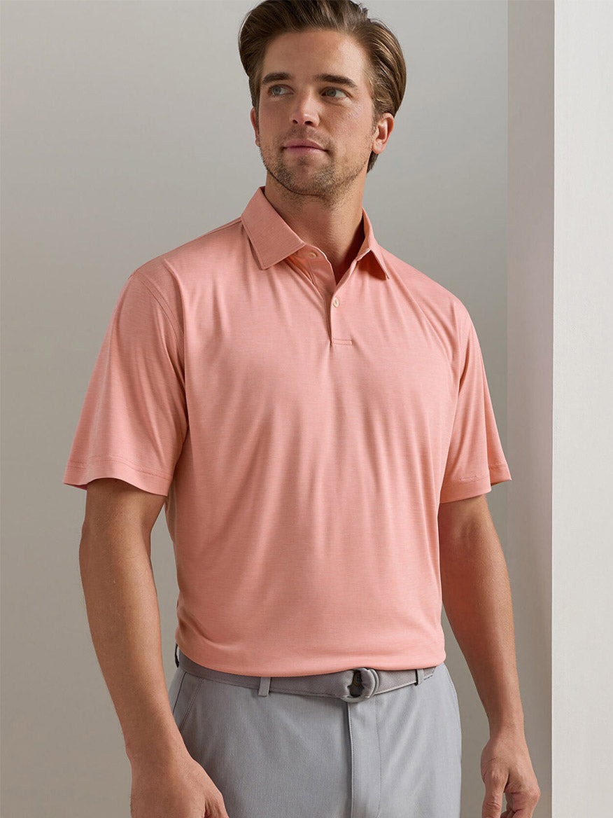 Man wears a Peter Millar Featherweight Mélange Performance Polo in Venetian Pink with UPF 50+ and light gray pants, standing indoors against a plain background.