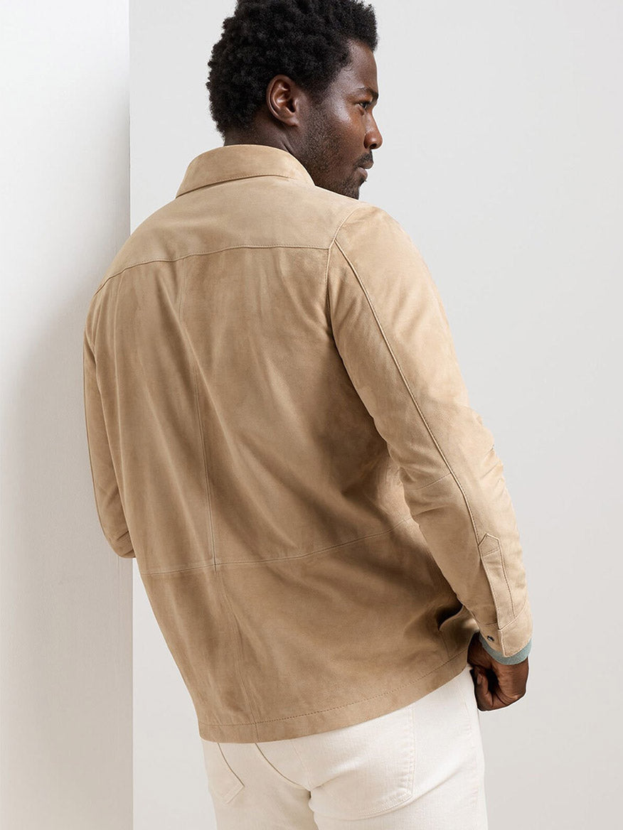 A man faces away, wearing the Peter Millar Featherweight Suede Shirt Jacket in Sand with off-white pants against a plain background.