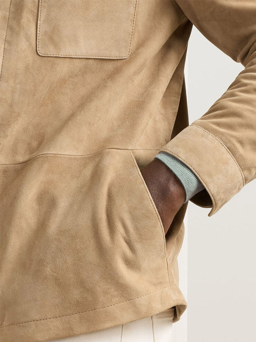 A person wearing the Peter Millar Featherweight Suede Shirt Jacket in Sand with a tailored fit and one hand in the pocket; part of a light blue sleeve and cream pants are also visible.