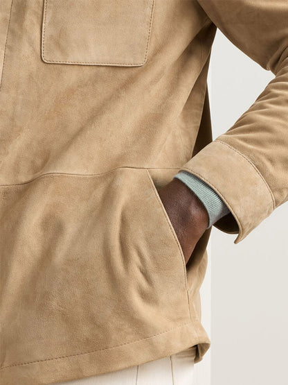 A person wearing the Peter Millar Featherweight Suede Shirt Jacket in Sand with a tailored fit and one hand in the pocket; part of a light blue sleeve and cream pants are also visible.