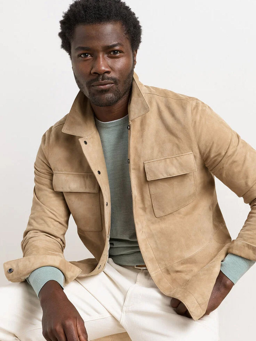 A man wearing the Peter Millar Featherweight Suede Shirt Jacket in Sand over a light green shirt and white pants is seated against a plain light background.
