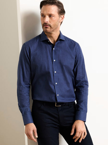 A man wearing the Peter Millar Felix Cotton Sport Shirt in Navy and dark pants stands against a white wall, looking to the side with a neutral expression.