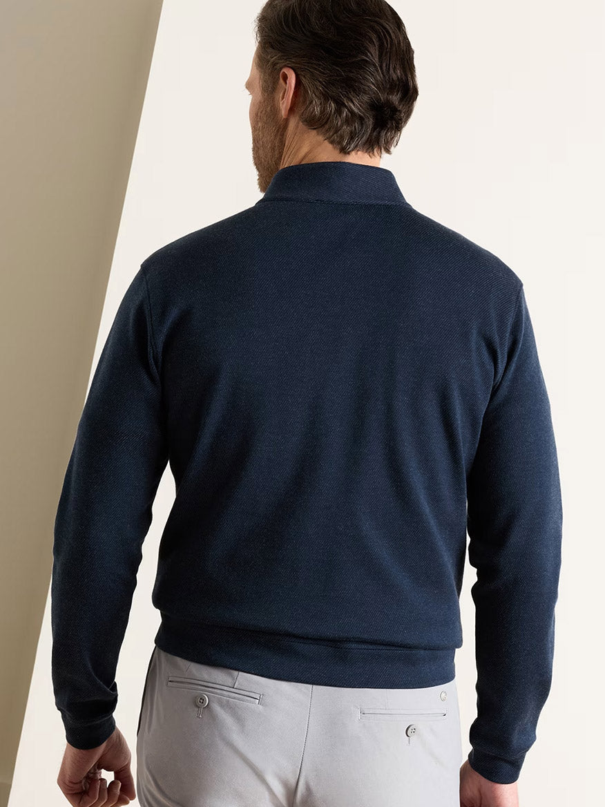 Man standing with his back to the camera, wearing a navy Peter Millar Grant Knit Quarter-Zip and light gray pants, against a neutral background.