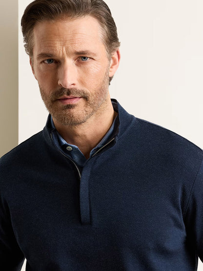 Man with light brown hair and beard wearing the Peter Millar Grant Knit Quarter-Zip in Navy, crafted from a soft cotton-cashmere blend, over a blue collared shirt, standing against an off-white background.