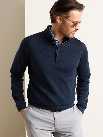 A man wearing sunglasses, a navy Peter Millar Grant Knit Quarter-Zip, and light gray pants stands with one hand in his pocket, looking to the side.