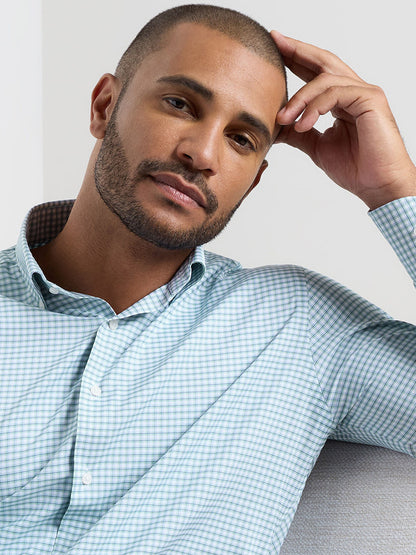 A man with a trimmed beard and short hair sits on a light-colored couch, wearing the Peter Millar Gregory Performance Twill Sport Shirt in Windsor Blue, and rests his head on his hand.