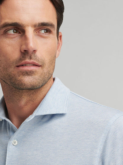 A man with short dark hair looks to the side, wearing a Peter Millar Harris Herringbone Knit Polo in Blue Frost tailored fit, against a plain gray background.