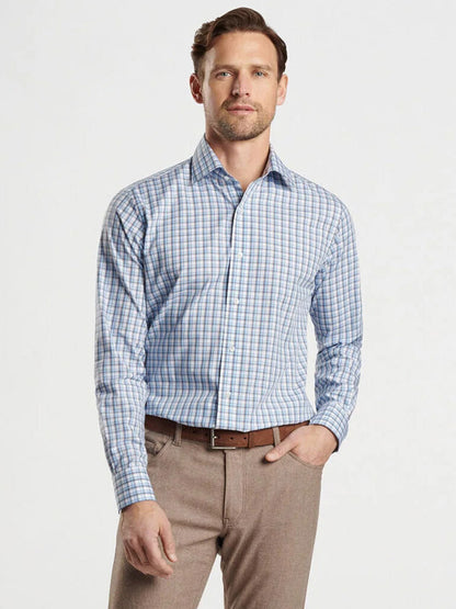 A man in the Peter Millar Howgill Cotton Sport Shirt in Brook Blue, crafted from premium cotton yarn, and beige pants with a tailored fit, stands with one hand in his pocket against a plain background.