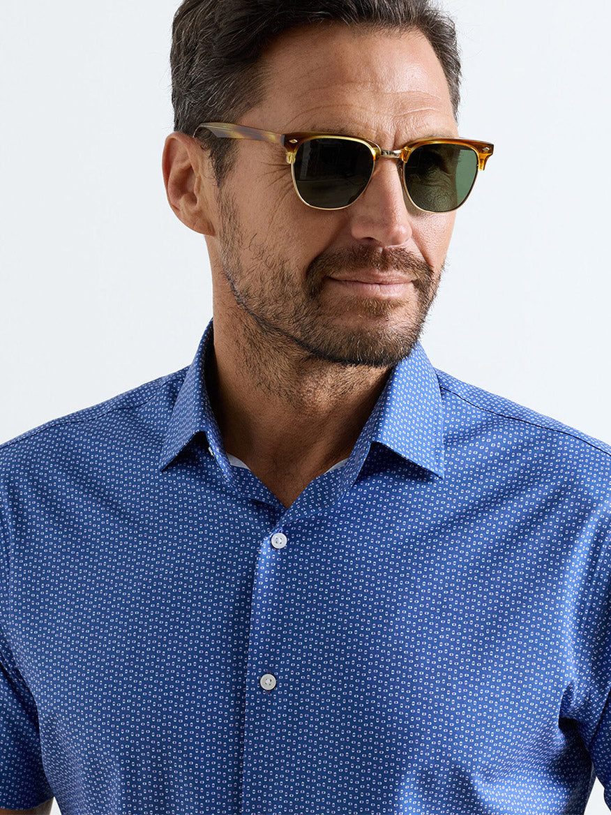 Man with short dark hair and beard wearing Peter Millar tortoiseshell sunglasses and the Jackson Performance Poplin Sport Shirt in Windsor Blue with UPF 50+, looking to the side against a plain background.