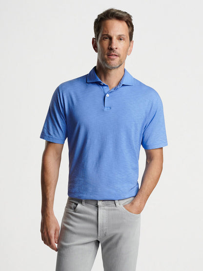 A man wearing a Peter Millar Journeyman Short Sleeve Polo in Regatta Blue and gray trousers standing against a neutral background.