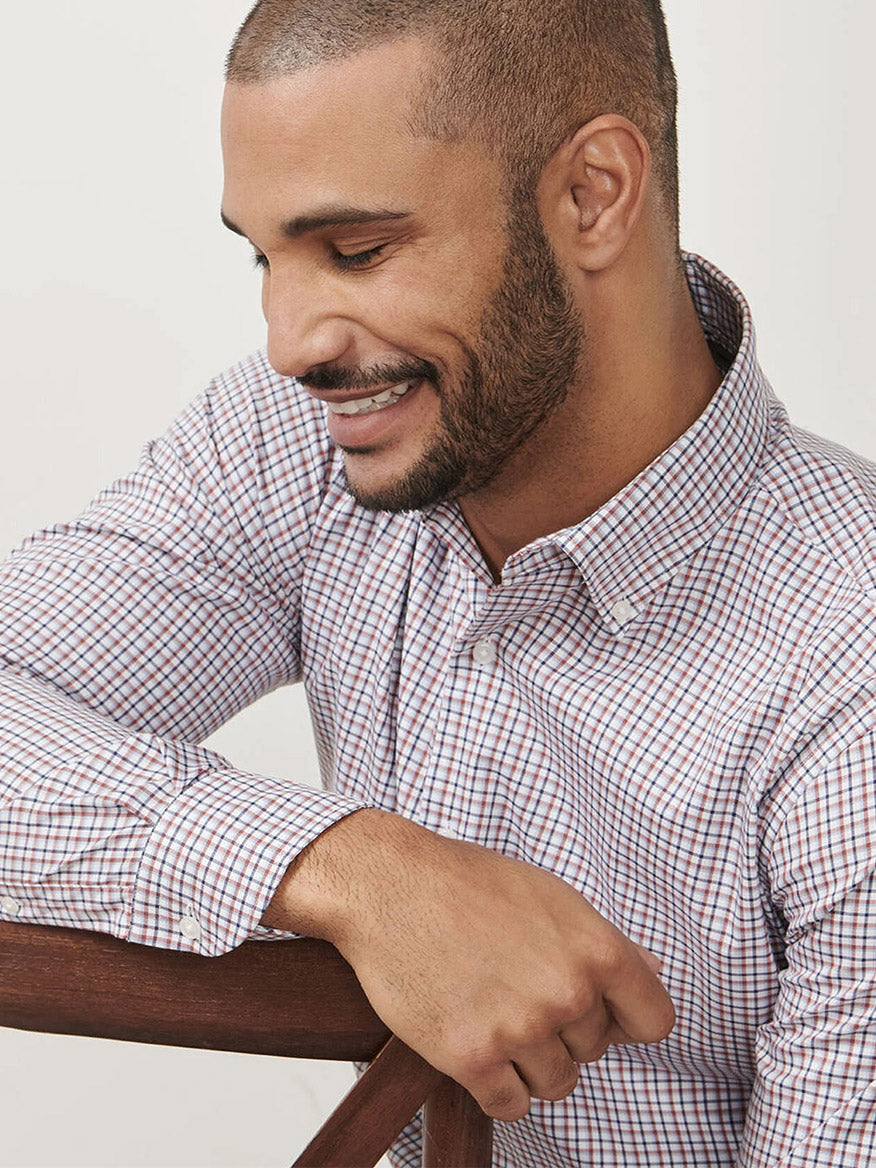 A man with short hair and a beard, smiling as he looks down while sitting sideways on a wooden chair, wears the Peter Millar Julian Crown Lite Cotton-Stretch Shirt in Navy.