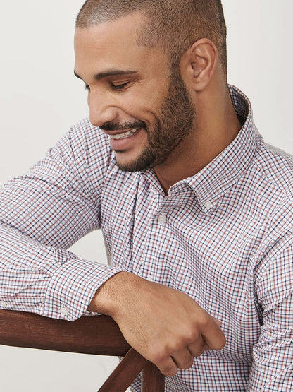 A man with short hair and a beard, smiling as he looks down while sitting sideways on a wooden chair, wears the Peter Millar Julian Crown Lite Cotton-Stretch Shirt in Navy.