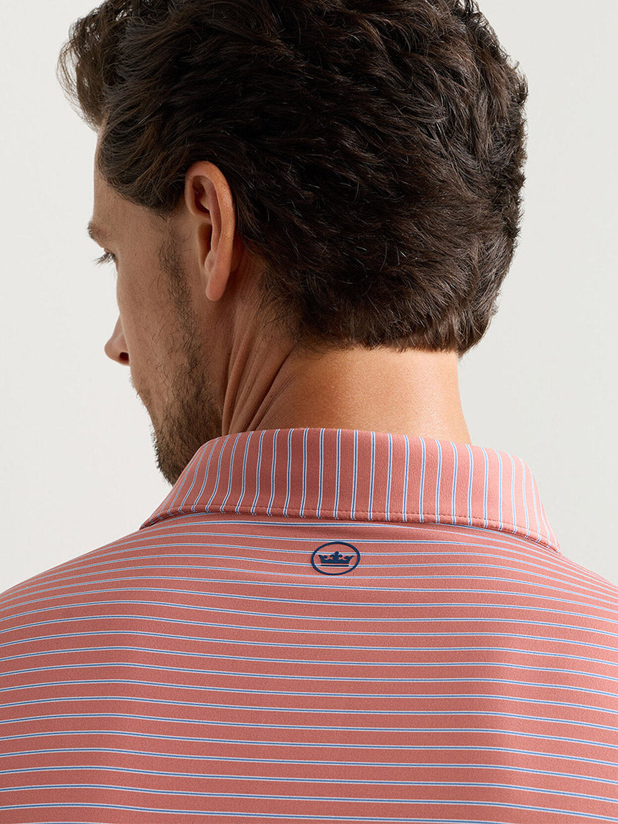 A man is seen from behind wearing the Peter Millar Keynote Performance Jersey Polo in Faded Scarlet, featuring white horizontal stripes, four-way stretch fabric, and a small crown logo at the back of the collar.